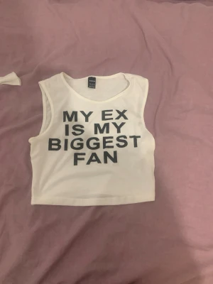 Vit croppad linnetopp med text - Säljer en vit croppad linnetopp från Shein med svart tryck 'MY EX IS MY BIGGEST FAN' på framsidan. Toppen är ärmlös och har en avslappnad passform, perfekt för att göra ett statement.
