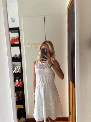 Vit spetsklänning med ärmlös design - Säljer en vit ärmlös klänning med vacker spets och rund halsringning. Klänningen har en medellång längd och ett lätt utställt fall. Perfekt för sommarens alla tillfällen. 