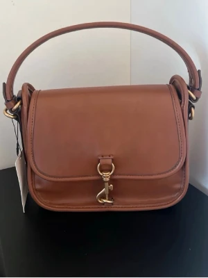 Brun axelremsväska från Zara  - Snygg brun handväska från Zara i slätt läder med guldfärgade metalldetaljer och ett dekorativt spänne framtill. Väskan har en stadig axelrem och klassisk, rektangulär form. Perfekt för dig som gillar stilrena accessoarer med exklusiv känsla.