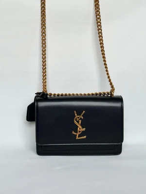 Svart YSL handväska med guldkedja - Snygg svart handväska från Saint Laurent med klassisk YSL-logga i guld på framsidan. Väskan har en stilren rektangulär form, guldfärgad kedjerem och detaljer i svart läder. Perfekt accessoar för att lyfta vilken outfit som helst.