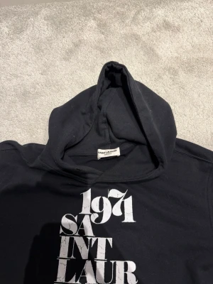 Svart hoodie från Saint Laurent - Säljer en svart hoodie från Saint Laurent med vit tryckt text '1971 SAINT LAURENT' på framsidan. Hoodien har klassisk huva och ribbade muddar vid ärmsluten. Tillverkad i mjukt bomullsmaterial som känns skönt mot huden. Perfekt för en avslappnad och trendig look.