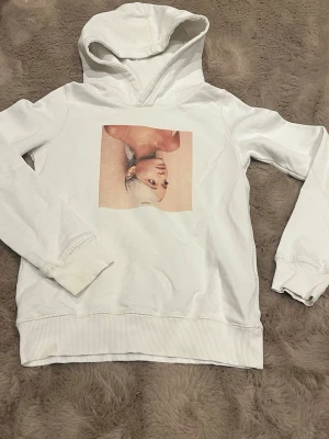 Vit hoodie med Sweetener-tryck - Vit hoodie med huva och tryck av albumomslaget till Ariana Grandes 'Sweetener' på bröstet. Hoodien har ribbade muddar vid ärmslut och nederkant. Perfekt för dig som gillar stilrena plagg med musiktema.