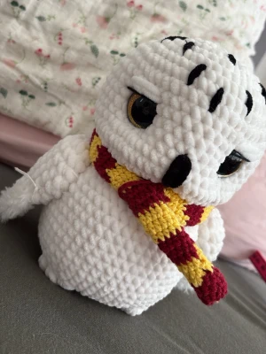 Virkad uggla med Gryffindor-halsduk - Söt handvirkad uggla i mjukt vitt garn med stora svarta och gula ögon. Ugglan har broderade svarta detaljer och bär en stickad halsduk i rött och gult, inspirerad av Gryffindor från Harry Potter. Perfekt som dekoration eller present till HP-fans. Ugglan är 20cm hög