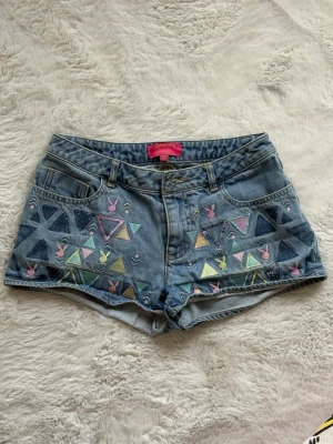 Playboy denimshorts med pastellmönster - Snygga denimshorts från Playboy med broderade pastellfärgade trianglar och små rosa Playboy-kaniner. Klassisk femficksmodell med knapp och dragkedja. 