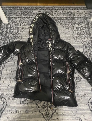 Svart dunjacka från Moncler med huva - Snygg svart dunjacka från Moncler med glansigt material och stor huva. Jackan har dragkedjor med detaljer i rött, vitt och blått samt Moncler-logga på huvan och ärmen. Två framfickor med dragkedja och puffig passform för extra värme.