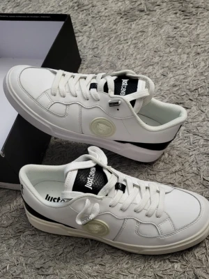 Vita Just Cavalli sneakers - Oanvända sneakers från Just Cavalli med svart detalj på hälen och svart logga på plösen. Skorna har en låg profil, vita snören och är tillverkade i slätt läder. Sidan pryds av en diskret logotyp i vitt. Perfekta för en clean och modern stil.
