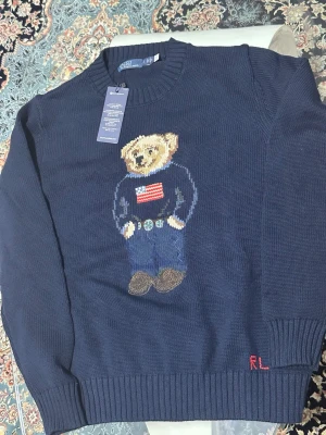 Mörkblå stickad tröja Polo Bear - Mörkblå stickad tröja från Polo Ralph Lauren med den ikoniska Polo Bear-björnen på bröstet, klädd i jeans och tröja med amerikansk flagga. Rund halsringning och ribbade muddar. RL-broderi i rött vid nederkanten. Perfekt för dig som gillar klassisk preppy stil.