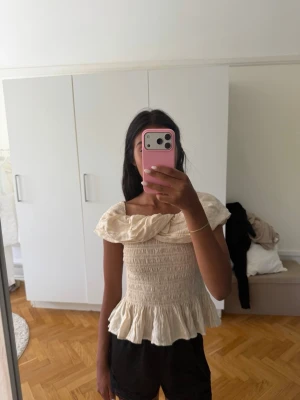 Beige offshoulder blus med volang - Säljer en söt beige offshoulder blus med bred volang runt axlarna och smockad midja. Toppen har en luftig och flowy känsla med volangkant nedtill, perfekt för varma dagar. Materialet känns lätt och bekvämt. Använd endast en gång!😊
