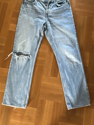 Ljusblå raka jeans med hål - Ljusblå jeans med rak passform och slitning samt stort hål på vänster knä. Klassisk femficksmodell med dragkedja och knapp. Jeansen har en avslappnad vibe och är tillverkade i denim, perfekt för en cool streetstil.