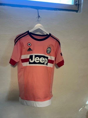 Juventus rosa matchtröja Adidas - Juventus rosa matchtröja från Adidas med svarta och vita detaljer, tre svarta ränder på axlarna och Jeep-logga på bröstet. Juventus klubbmärke med tre stjärnor och rund hals. Tillverkad i lätt och ventilerande material.