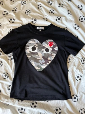Svart Comme des Garçons PLAY t-shirt - Svart t-shirt från Comme des Garçons PLAY med ett stort hjärta i camouflage på bröstet och en liten röd hjärtdetalj. Klassisk rund hals och korta ärmar. Perfekt statement-plagg för dig som gillar streetwear och coola prints.