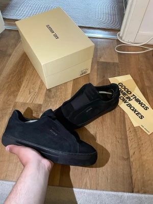 Svarta sneakers från Axel Arigato - Stilrena svarta sneakers från Axel Arigato i mocka med minimalistisk design och diskret logga på sidan. Skorna har en låg siluett och svart sula som ger en clean look. Perfekta för dig som gillar enkel och modern stil.