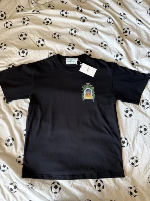 Svart Casablanca t-shirt med tryck - Svart t-shirt från Casablanca med färgglatt tryck av ett fönster och gröna blad på ryggen samt mindre tryck på bröstet. Klassisk passform och rund hals. Perfekt för dig som vill ha en unik och stilren tee med snygga detaljer.