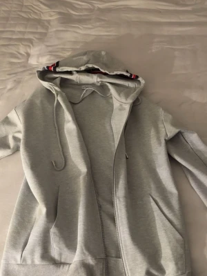 Grå Moncler hoodie med dragkedja - Snygg grå hoodie från Moncler med dragkedja, huva med dragsko och ribbade muddar med Monclers signaturfärger. Klassisk logga på ärmen och fickor framtill. Perfekt för en chill och stilren look. Aldrig använd då den ej passar min storlek!