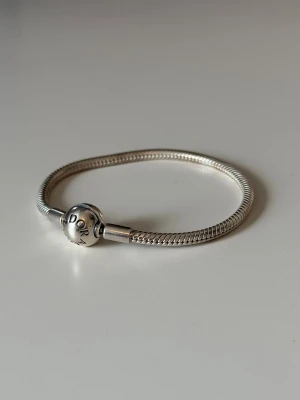 Pandora silverarmband med pärldetalj - Snyggt silverfärgat armband från Pandora med en rund pärldetalj som lås, där Pandora-loggan är ingraverad. Armbandet har en klassisk och stilren design i metall som passar perfekt för att addera egna berlocker. 