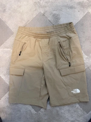 Beige shorts från The North Face - Snygga beige shorts från The North Face med elastisk midja och två stora cargofickor. Shortsen har dragkedjor på sidofickorna och logga på vänster ben. Perfekta för sommardagar och outdoor-äventyr.