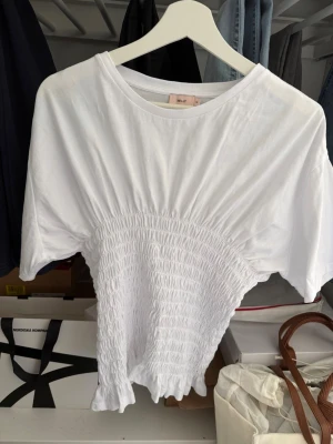 Vit topp med smock från Nelly - Säljer en vit topp från Nelly med smockad midja och korta ärmar. Toppen har rund halsringning och är gjord i mjuk bomull. Perfekt för en avslappnad och trendig look.