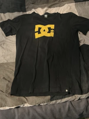 Svart DC t-shirt med gul logga - Säljer en svart t-shirt från DC med stor gul logga på bröstet. Klassisk passform och rund halsringning. Perfekt för dig som gillar streetwear och vill ha en enkel men snygg tee. Materialet känns som bomull och t-shirten har korta ärmar.