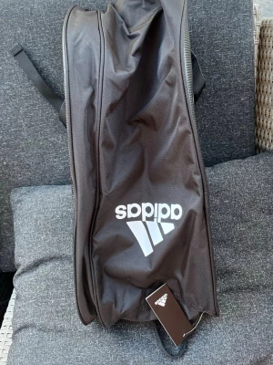 Svart padelväska från Adidas - Hej!  Säljer en svart padelväska från Adidas. Perfekt för träning eller att bära sportutrustning.  Vill du veta något mer eller vill ha fler bilder så är det bara att skriva! Priset är såklart inte hugget i sten!