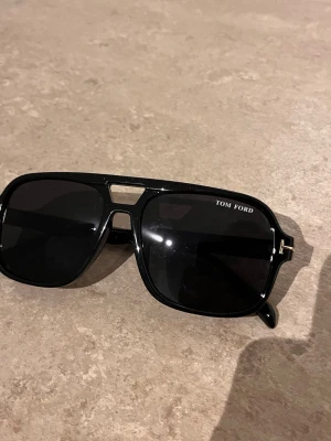 Svarta Tom Ford pilotbågar solglasögon - Snygga svarta solglasögon från Tom Ford med klassisk pilotform och bred båge. Glasögonen har mörka linser och diskreta gulddetaljer vid skalmarna. Perfekta för dig som vill ha en stilren och exklusiv look.