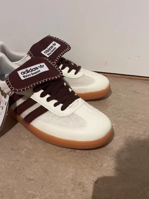 Adidas Wales Bonner sneakers brun/vit - Snygga Adidas Wales Bonner sneakers med brun mockadetalj, vita läderpaneler och klassiska tre ränder i brunt. Tjock gummisula och unik oversized plös med kontrastsöm. Perfekt för dig som gillar retrovibbar och streetstyle.