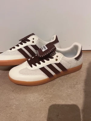 Adidas Wales Bonner sneakers brun/vit - Säljer ett par Adidas Wales Bonner sneakers med brun mocka och vita detaljer. Skorna har klassiska tre ränder i brunt, vit ovandel i läder och mocka, samt gummisula. Tungan är extra stor med dekorativ söm och Wales Bonner-logga. Perfekta för dig som gillar retrostil.