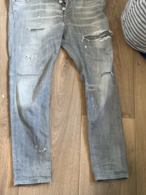 Grå slitna jeans från Dsquared2 - Säljer ett par grå jeans från Dsquared2 med skinny fit, slitningar och hål på benen samt vita färgstänk för en cool, trendig look. Jeansen har klassisk femficksmodell och knappgylf. Perfekta för dig som gillar streetstyle och edgy mode.