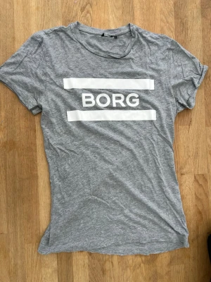 Grå t-shirt från BORG - Snygg grå t-shirt från BORG med vit logga framtill. Klassisk passform med rund hals och korta ärmar. Tillverkad i mjukt bomullsmaterial som känns skönt mot huden. Perfekt till jeans eller shorts för en avslappnad look.