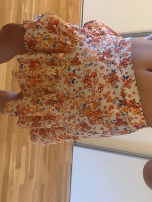 Blommig volangkjol i orange och vit - Supersöt kort kjol med volanger och blommigt mönster i orange, blått och vitt. Kjolen har en luftig passform och är perfekt för varma dagar. Tillverkad i ett lätt och mjukt material som känns skönt mot huden.