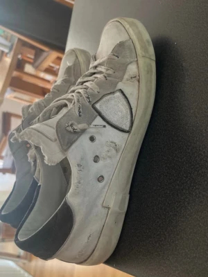Philippe Model vita sneakers - Säljer ett par vita Philippe Model sneakers med slitna detaljer och svart hälparti. Skorna har låga skaft, klassisk snörning och är tillverkade i läder och textil. Ikonisk sköldlogga på sidan och fransade kanter för en edgy look.