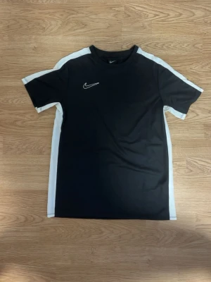 Svart Nike t-shirt med vita detaljer - Snygg svart t-shirt från Nike med vita paneler längs sidorna och ärmarna. Klassisk rund halsringning och korta ärmar. Passar dig som är mellan 160-170 cm lång. Perfekt för en sportig look och tillverkad i mjukt, lätt material som andas.