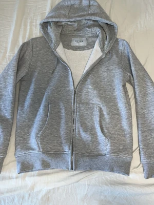 Grå RYVLS Stockholm hoodie  - Mysig grå hoodie från RYVLS Stockholm med dragkedja, huva och två stora fickor framtill. Insidan är fodrad med mjukt, vitt teddyfoder som gör den extra skön. Perfekt för dig som gillar en enkel och stilren look. Använd 2 gånger och tvättad. Helt nya utan skador.