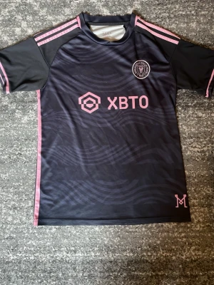 Inter Miami svart fotbollströja - Snygg svart fotbollströja från Adidas för Inter Miami CF med rosa detaljer och klubbmärke på bröstet. Tröjan har korta ärmar, rund hals och rosa ränder på axlarna med messi numer 10 på ryggen. Tillverkad i lätt och ventilerande material, perfekt för träning eller match mycket bra skick. storlek m