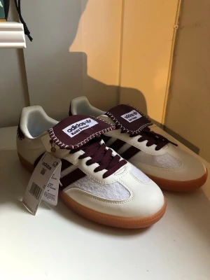 Adidas Wales bonner vit/vinröd sneaker - Adidas Samba OG sneakers i vitt och vinrött med klassiska tre ränder på sidan, brun gummisula och detaljerad kontrastsöm på plösen. Ovandelen är i läder och mesh, med vinröda skosnören och Adidas-logga på plösen. Perfekt för dig som gillar retrostil.