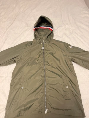 Olivgrön vindjacka från Moncler 💚 - Snygg olivgrön vindjacka från Moncler med luva och dragkedja framtill. Jackan har två fickor med tryckknappar och en Moncler-logga på ärmen. Luvan har en cool detalj med röd, vit och blå rand. Tillverkad i lätt, glansigt material.                                                                           OBS: om du är vid 190+ och väger över 90 kommer denna produkt passa dig!! 