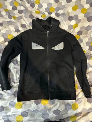 Svart hoodie med ögon från Fendi - Cool svart hoodie från Fendi med dragkedja och huva. På bröstet finns två silvriga ögon som ger en unik och edgy look. Hoodien har två fickor framtill och ribbade muddar vid ärmslut och nederkant. Perfekt för dig som vill sticka ut.