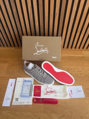 Gråa Christian Louboutin sneakers - Snygga gråa sneakers från Christian Louboutin med klassisk röd sula och vita kanter. Skorna är låga och har en stilren design i mocka med matchande grå skosnören. Perfekta för dig som vill ha något exklusivt och trendigt. Storlek 42! Och aldrig använda! Vid frågor eller funderingar är de bara att höra av sig!