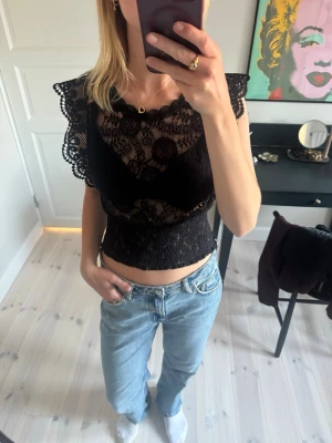 Svart spetsblus med volangdetaljer - Supersnygg svart blus i spets med volangdetaljer på axlarna. Har en stretchig resår i midjan vilket gör att den funkar om man oftast bär en storlek större eller mindre. I fint skick.
