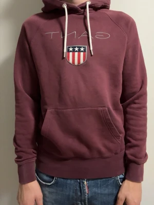 Vinröd hoodie från Gant med tryck - Säljer en vinröd hoodie från Gant med vit dragsko i huvan och ett broderat Gant-emblem med stjärnor och ränder på bröstet. Hoodien har känguruficka framtill och ribbade muddar vid ärmar och nederkant. Perfekt för en avslappnad och snygg stil.