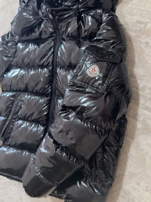 Svart glansig dunjacka från Moncler - Snygg svart dunjacka från Moncler med glansig finish, huva och dragkedja framtill. Jackan har två snedställda fickor med dragkedja och Moncler-logga på ärmen. Perfekt för kalla vinterdagar och riktigt trendig look. För mer info kontakta mig 