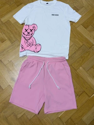 Set med vit t-shirt och rosa shorts - Säljer ett set med en vit t-shirt med trycket 'NEVER' och en stor rosa nalle med kedja på framsidan. Tillhörande rosa shorts med dragsko och bakficka. Materialet ser ut att vara mjukt och bekvämt, perfekt för en avslappnad stil.