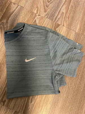 Grå Nike Dri-Fit t-shirt - Snygg grå Nike Dri-Fit t-shirt med diskret mönster och klassisk logga på bröstet. Perfekt för löpning eller träning, tillverkad i ett lätt och snabbtorkande syntetmaterial. Rund halsringning och korta ärmar för en sportig look.
