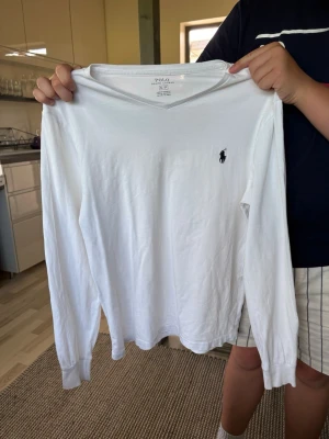 Vit långärmad tröja Polo Ralph Lauren - Klassisk vit långärmad tröja från Polo Ralph Lauren med V-ringning och den ikoniska svarta logobroderingen på bröstet. Tillverkad i mjuk bomull som känns skön mot huden. Perfekt till jeans eller shorts för en clean och stilren look.