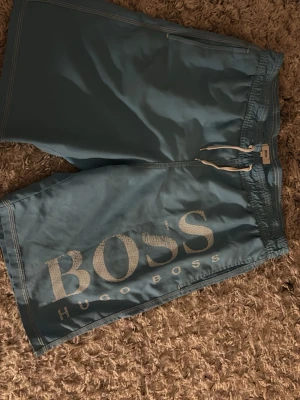 Blå badbyxor från Hugo Boss - Snygga blå badbyxor från Hugo Boss med stort vitt BOSS-tryck på ena benet. Klassisk modell med resår i midjan och vit dragsko. Perfekta för stranden eller poolen, tillverkade i snabbtorkande material.