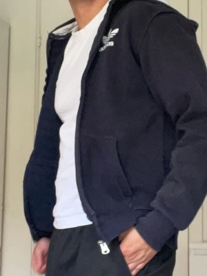 Marinblå / Svart ziptröja från Adidas - Klassisk svart / marinblå zip up hoodie från Adidas med dragkedja framtill och vit logga på bröstet. Tröjan har huva med dragsko och två fickor fram. Tillverkad i mjukt sweatshirtmaterial som är skönt att bära. Perfekt för en avslappnad och sportig stil.