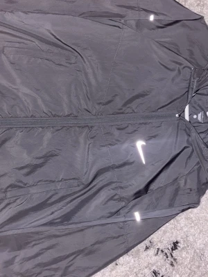 Nike Miller  - Snygg svart vindjacka från Nike med dragkedja framtill och två sidofickor. Jackan har vita reflexdetaljer och Nike-logga på bröstet. Tillverkad i lätt och slitstarkt material, perfekt för löpning eller andra utomhusaktiviteter.