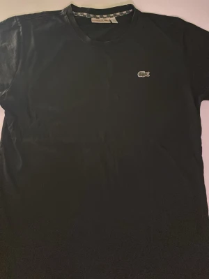 Svart Lacoste t-shirt  - Klassisk svart t-shirt från Lacoste med rund hals och den ikoniska krokodilloggan broderad på bröstet. Modellen är enkel och stilren, tillverkad i mjuk bomull och har en normal passform. Perfekt för en clean och avslappnad look.