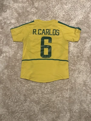 Brasilien fotbollströja R. Carlos Nike - Gul Brasilien fotbollströja från Nike med gröna detaljer, nummer 6 och R. Carlos på ryggen. Tröjan har rund hals, korta ärmar och Brasilien-emblem på bröstet. Perfekt för dig som älskar fotboll och vill bära en ikonisk landslagströja.