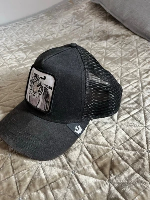Svart truckerkeps från goorin bros  - Säljer en svart truckerkeps från Goorin Bros med mesh på sidorna och bak. Framtill finns ett tygmärke med en tiger. Justerbar snapback och böjd skärm. 