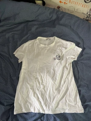 Vit Moncler t-shirt med logga - Säljer en vit t-shirt från Moncler med klassisk rund hals och korta ärmar. På bröstet sitter två snygga Moncler-loggor i blått och rött. Perfekt basic-plagg med exklusiv känsla och skön passform i bomull.